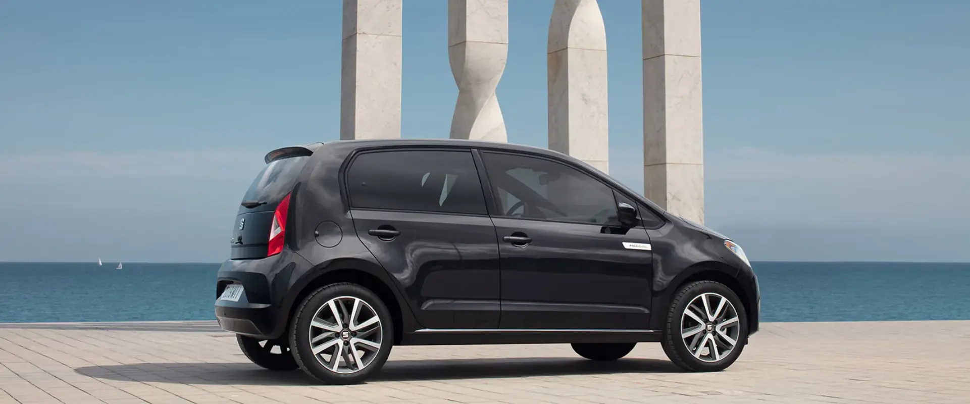 Sort SEAT Mii-bil parkeret ved en havnepromenade med skulpturelle søjler og havet i baggrunden.