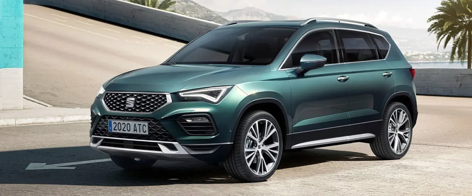 En turkisgrøn SEAT Ateca SUV holder parkeret på en solrig kystvej med bjerge i baggrunden.