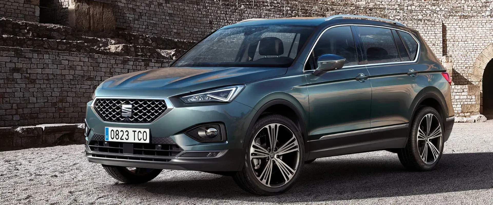 En blågrøn SEAT Tarraco SUV holder parkeret foran en gammel stenmur i sollys.